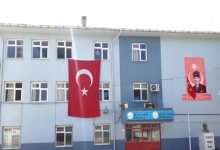 Mavi-beyaz boyalı bir okul binasının cephesinde büyük bir Türk bayrağı ve Atatürk posteri asılı. Girişte “Refik Pınar İlkokulu” ve “Refik Pınar Ortaokulu” tabelaları bulunuyor.