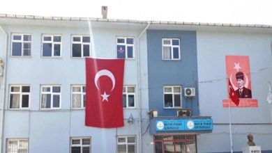 Mavi-beyaz boyalı bir okul binasının cephesinde büyük bir Türk bayrağı ve Atatürk posteri asılı. Girişte “Refik Pınar İlkokulu” ve “Refik Pınar Ortaokulu” tabelaları bulunuyor.