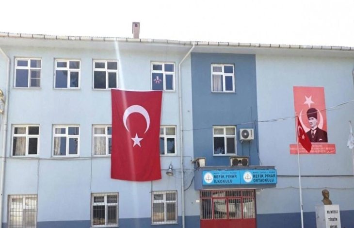 Mavi-beyaz boyalı bir okul binasının cephesinde büyük bir Türk bayrağı ve Atatürk posteri asılı. Girişte “Refik Pınar İlkokulu” ve “Refik Pınar Ortaokulu” tabelaları bulunuyor.
