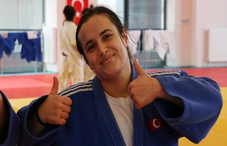 Görselde, mavi judogi giymiş bir kadın sporcu kameraya gülümseyerek iki başparmağını yukarı kaldırıyor. Arka planda antrenman salonu ve diğer judogiler görünüyor. Sporcu enerjik ve gururlu bir ifade taşıyor.