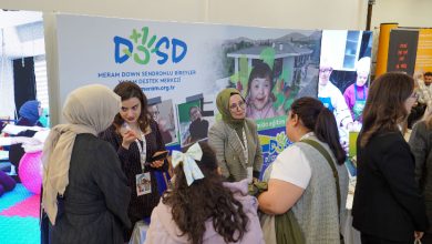 Görselde, bir fuar ya da tanıtım etkinliğinde “Meram Down Sendromlu Bireyler Yaşam Destek Merkezi (DOSD Meram)” standı görülüyor. Standın önünde bir grup kadın sohbet ediyor. Arka planda merkezin logosu, web adresi ve down sendromlu bireylerin yer aldığı fotoğraflar yer alıyor. Ortam canlı ve bilgilendirici bir etkileşim atmosferinde.