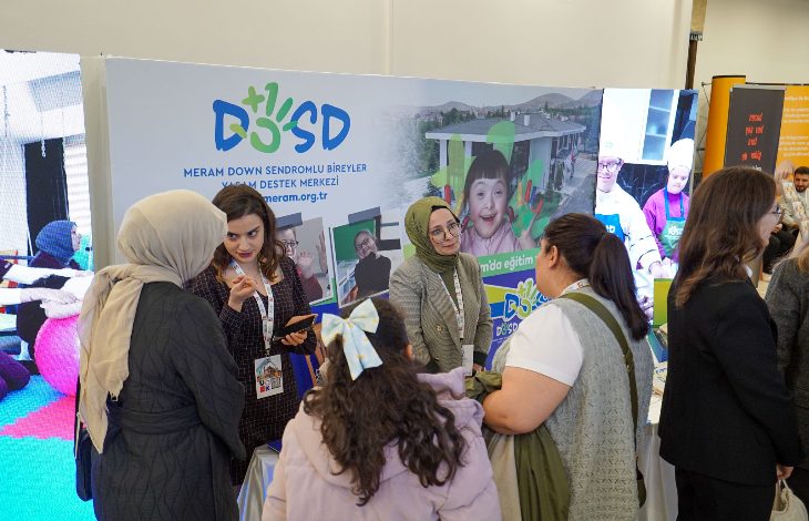 Görselde, bir fuar ya da tanıtım etkinliğinde “Meram Down Sendromlu Bireyler Yaşam Destek Merkezi (DOSD Meram)” standı görülüyor. Standın önünde bir grup kadın sohbet ediyor. Arka planda merkezin logosu, web adresi ve down sendromlu bireylerin yer aldığı fotoğraflar yer alıyor. Ortam canlı ve bilgilendirici bir etkileşim atmosferinde.
