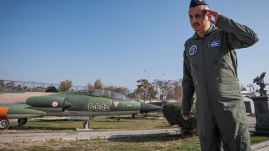 Görselde, askerî pilot tulumu giymiş bir erkek, açık havada selam dururken görülüyor. Üzerinde arma ve isim etiketi bulunan yeşil uçuş tulumu var, başında mavi bir pilot kepi taşıyor. Arka planda ise açık alanda sergilenen, üzerinde “8-642” numarası bulunan bir savaş uçağı duruyor. Hava açık, ortam bir hava müzesi ya da askeri sergi alanını andırıyor.
