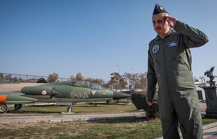 Görselde, askerî pilot tulumu giymiş bir erkek, açık havada selam dururken görülüyor. Üzerinde arma ve isim etiketi bulunan yeşil uçuş tulumu var, başında mavi bir pilot kepi taşıyor. Arka planda ise açık alanda sergilenen, üzerinde “8-642” numarası bulunan bir savaş uçağı duruyor. Hava açık, ortam bir hava müzesi ya da askeri sergi alanını andırıyor.