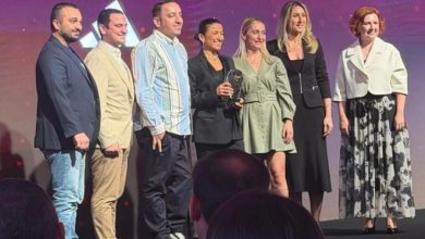 Bir sahnede yan yana dizilmiş yedi kişilik bir grup görülüyor. Hepsi şık ve modern kıyafetlerle poz veriyor. Ortadaki iki kişi küçük, cam görünümlü bir ödül objesini birlikte tutuyor. Arka planda mor ve pembe tonlarda ışıklı bir sahne efekti var. Ön planda ise izleyici siluetleri seçiliyor. Tören atmosferi belirgin.