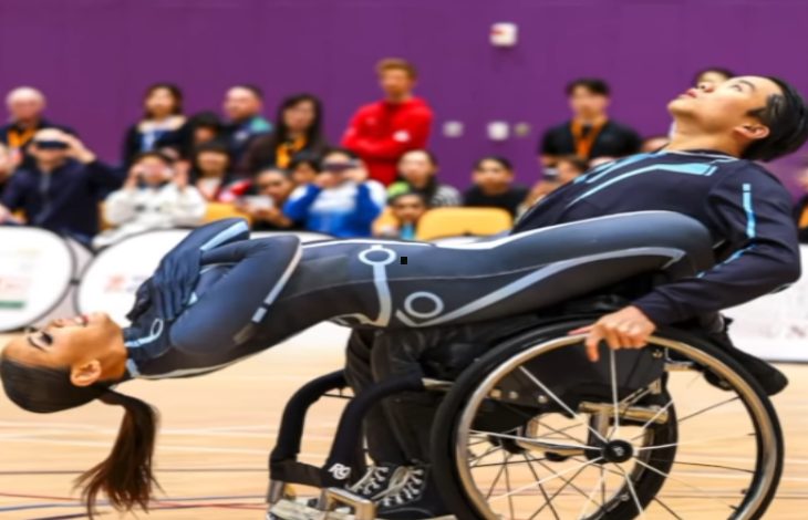 Bir spor salonunda, tekerlekli sandalyedeki bir dansçı partneriyle birlikte etkileyici bir dans figürü sergiliyor. Arkada izleyici topluluğu sahneyi dikkatle izliyor.