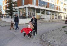 Görselde, tekerlekli sandalyede oturan küçük bir kız çocuğu bulunuyor. Üstü kırmızı bir battaniyeyle örtülü. Sandalyeyi başörtülü bir kadın itiyor. Arkada, bir erkek ve yanında yürüyen küçük bir çocuk yer alıyor. Hepsi bir apartman ve iş yerlerinin bulunduğu bir sokakta ilerliyor.