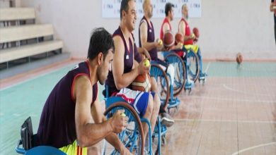 Bir spor salonunda, tekerlekli sandalyede oturan erkek sporcular basketbol toplarıyla antrenman yapıyor. Oyuncular sıralı şekilde dizilmiş ve odaklanmış görünüyor.
