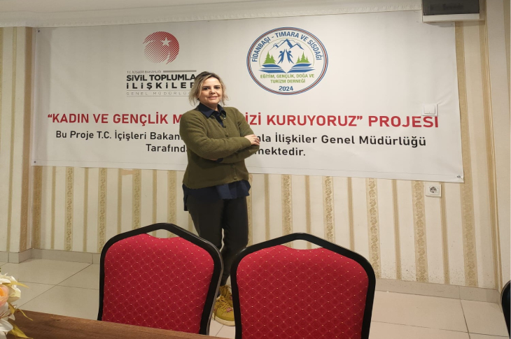 Görselde, bir kadın ayakta durarak kameraya gülümseyen bir poz veriyor. Arkasında, “Kadın ve Gençlik Merkezimizi Kuruyoruz” başlıklı proje afişi yer alıyor. Önde kırmızı sandalyeler ve bir masa görülüyor. Ortam bir toplantı ya da dernek salonunu andırıyor.