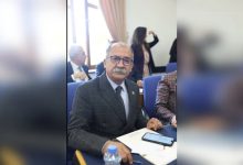 Görselde takım elbiseli, gözlüklü, bıyıklı bir erkek bir toplantı salonunda masada oturuyor. Önünde notlar ve telefonu var. Arkada başka katılımcılar da görünüyor.