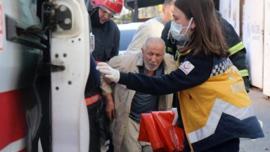 Bir acil durum sahnesi var. Bir ambulansın kapısı açık, sağlık görevlisi maske takmış hâlde yaşlı bir adamı araca yönlendiriyor. Yanlarında itfaiyeciler de bulunuyor; üzerlerinde koruyucu ekipmanlar var. Adam yorgun ve tedirgin görünüyor, ekip ise hızlı ve dikkatli şekilde müdahale ediyor. Ortamda yoğun bir hareketlilik ve acil yardım havası hissediliyor.