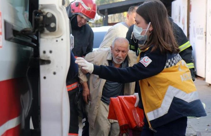 Bir acil durum sahnesi var. Bir ambulansın kapısı açık, sağlık görevlisi maske takmış hâlde yaşlı bir adamı araca yönlendiriyor. Yanlarında itfaiyeciler de bulunuyor; üzerlerinde koruyucu ekipmanlar var. Adam yorgun ve tedirgin görünüyor, ekip ise hızlı ve dikkatli şekilde müdahale ediyor. Ortamda yoğun bir hareketlilik ve acil yardım havası hissediliyor.