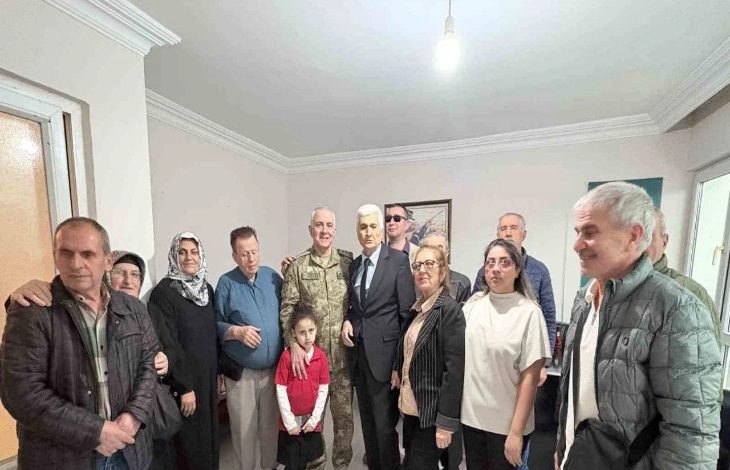 Fotoğrafta, bir araya gelmiş kalabalık bir grup görünüyor. Ortada üniformalı bir asker ile takım elbiseli bir kişi yan yana duruyor; etraflarında farklı yaşlardan kadınlar, erkekler ve bir çocuk bulunuyor. Herkes kameraya dönük, samimi ve sıcak bir aile buluşması havası var. Oda sade, duvarda bir tablo ve tavanda tek bir ampul ışığı görülüyor.