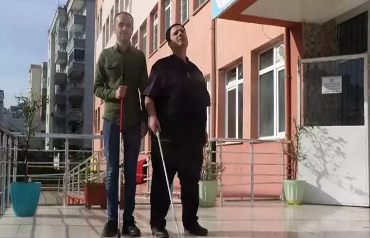 Fotoğrafta, bir okul binasının girişinde iki görme engelli öğretmen yan yana duruyor. Her ikisi de beyaz bastonlarıyla ayakta ve kendinden emin bir duruş sergiliyor. Soldaki öğretmen ince yapılı, yeşil bir gömlek giymiş; sağdaki öğretmen ise daha iri yapılı ve koyu renk bir kıyafet tercih etmiş. Arka planda okulun pembe tonlarındaki dış cephesi, pencereler ve giriş kapısı görünüyor. Güneşli bir hava, zemine düşen ışık ve gölgelerle belli oluyor. Sahne genel olarak güçlü, ilham veren bir birliktelik hissi taşıyor.