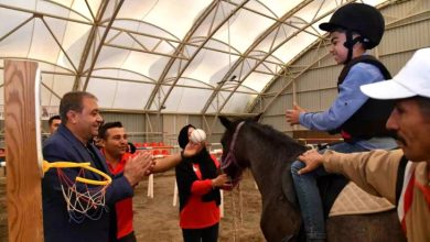 Kapalı manejde düzenlenen etkinlikte, bir çocuk at üzerinde terapi yaparken çevresindekiler onu destekleyip alkışlıyor. Çocuk kask takmış, gülümseyerek topa uzanıyor.