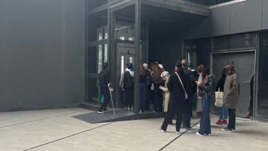 Bir grup insanın bir binanın girişindeki cam asansöre doğru sırayla ilerlediği, bazı kişilerin ise dışarıda bekleyerek telefonlarına baktığı görülüyor. Koltuk değneği kullanan bedensel engelli erkek ise yan tarafta bekliyor.