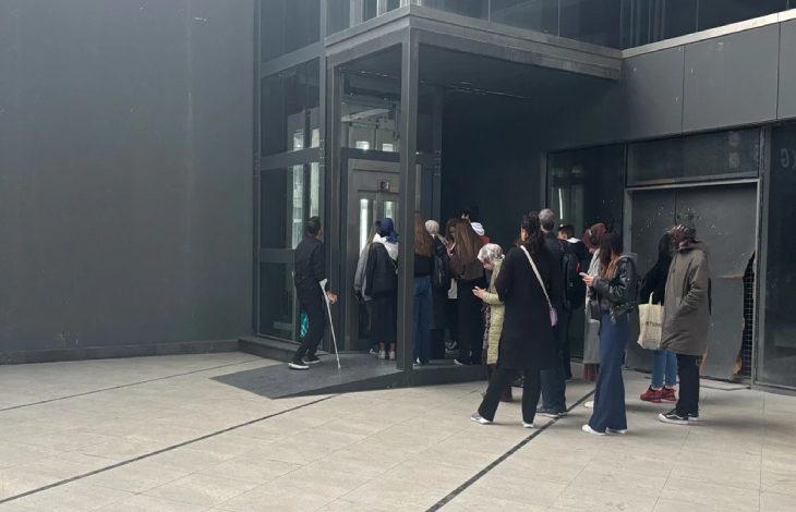 Bir grup insanın bir binanın girişindeki cam asansöre doğru sırayla ilerlediği, bazı kişilerin ise dışarıda bekleyerek telefonlarına baktığı görülüyor. Koltuk değneği kullanan bedensel engelli erkek ise yan tarafta bekliyor.