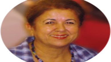 Fotoğrafta, yuvarlak çerçeve içinde orta yaşlarda bir kadın görünüyor. Sıcacık bir gülümsemeye sahip, yüz hatları yumuşak ve samimi. Kısa, koyu renk saçları geriye doğru taranmış. Üzerinde mavi tonlarında kareli bir gömlek var ve boynunda renkli boncuklardan oluşan bir kolye duruyor. Arka plan flu olduğu için dikkat tamamen kadının yüzüne ve ifadeye odaklanıyor; fotoğraf ona sakin, güven veren bir aura kazandırıyor.