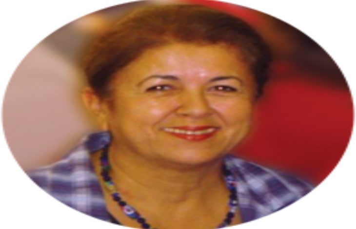 Fotoğrafta, yuvarlak çerçeve içinde orta yaşlarda bir kadın görünüyor. Sıcacık bir gülümsemeye sahip, yüz hatları yumuşak ve samimi. Kısa, koyu renk saçları geriye doğru taranmış. Üzerinde mavi tonlarında kareli bir gömlek var ve boynunda renkli boncuklardan oluşan bir kolye duruyor. Arka plan flu olduğu için dikkat tamamen kadının yüzüne ve ifadeye odaklanıyor; fotoğraf ona sakin, güven veren bir aura kazandırıyor.