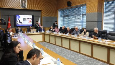 Bir toplantı salonunda uzun masa etrafında toplanmış kişiler görülüyor. Masada belgeler ve su şişeleri var, arka planda ise bir sunum ekranı ve Türk bayrağı yer alıyor.