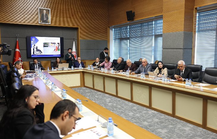 Bir toplantı salonunda uzun masa etrafında toplanmış kişiler görülüyor. Masada belgeler ve su şişeleri var, arka planda ise bir sunum ekranı ve Türk bayrağı yer alıyor.