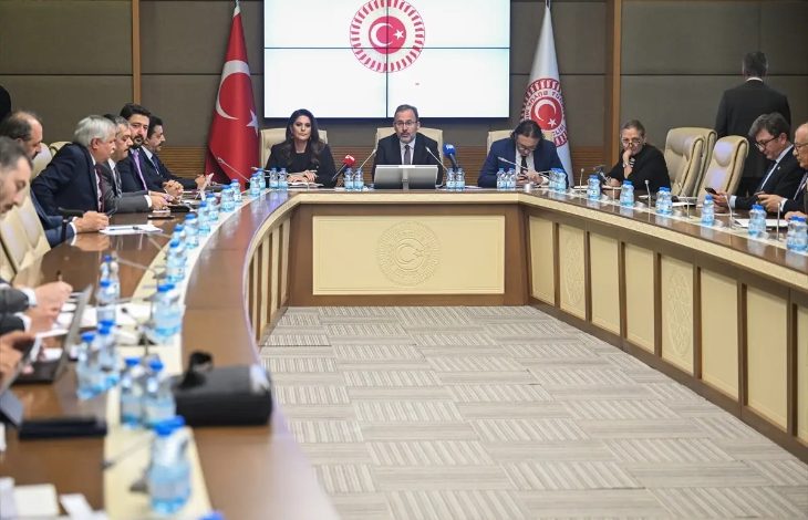 Görselde, Türkiye Büyük Millet Meclisi’nde gerçekleştirilen bir komisyon toplantısı görülüyor. Uzun, U şeklinde bir masanın etrafında birçok kişi oturmakta. Masanın ortasında mikrofonlar ve belgeler bulunuyor. Ortadaki ekranda ve masanın ön panelinde TBMM amblemi yer alıyor. Arka planda Türk bayrağı ve TBMM bayrağı dikkat çekiyor. Toplantı resmi ve ciddi bir atmosferde gerçekleşiyor.