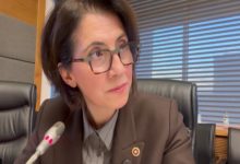 Gözlüklü bir kadın, mikrofonun önünde oturuyor ve dikkatle bir yöne bakıyor. Arka planda ofis ortamı ve büyük pencereler görünüyor.