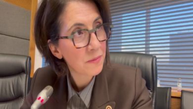 Gözlüklü bir kadın, mikrofonun önünde oturuyor ve dikkatle bir yöne bakıyor. Arka planda ofis ortamı ve büyük pencereler görünüyor.