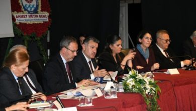 Bir salonda düzenlenen toplantıda, masada oturan kadın ve erkek katılımcılar mikrofonlarla konuşma yapıyor. Masada belgeler, mikrofonlar ve çiçek düzenlemeleri bulunuyor.