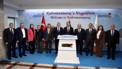 Bir grup yetkili ve görevli, “Kahramanmaraş’a Hoşgeldiniz” yazısının bulunduğu alanda yan yana dizilmiş şekilde poz veriyor. Ortada bir kürsü, arka planda Türk bayrağı ve belediye logosu görülüyor. Herkes resmi kıyafetleriyle kameraya bakıyor.