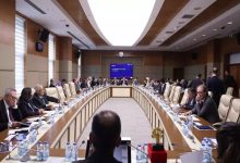 Geniş, modern bir toplantı salonunda uzun U şeklindeki masanın etrafını dolduran çok sayıda kişi görülüyor. Katılımcılar belgelerine eğilmiş, not alıyor ya da konuşmaları takip ediyor. Masaların üzerinde su şişeleri, dosyalar ve mikrofonlar yer alıyor. Salonun ortasında büyük bir ekran bulunuyor. Ortam, yoğun gündemli ve resmi bir komisyon toplantısı atmosferi taşıyor.