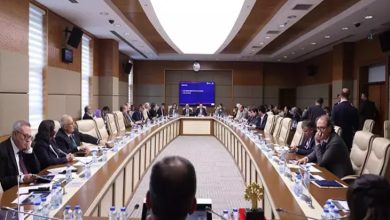 Geniş, modern bir toplantı salonunda uzun U şeklindeki masanın etrafını dolduran çok sayıda kişi görülüyor. Katılımcılar belgelerine eğilmiş, not alıyor ya da konuşmaları takip ediyor. Masaların üzerinde su şişeleri, dosyalar ve mikrofonlar yer alıyor. Salonun ortasında büyük bir ekran bulunuyor. Ortam, yoğun gündemli ve resmi bir komisyon toplantısı atmosferi taşıyor.