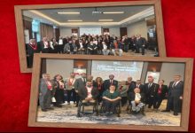 Kırmızı zemine yerleştirilmiş iki çerçeveli fotoğraf görünüyor. Üstteki karede geniş bir salonun içinde kalabalık bir grup birlikte poz veriyor; arka planda sunum perdesi açık. Alttaki karede yine aynı salonda, bu kez daha yakın planda bir grup fotoğrafı var. Ön sırada tekerlekli sandalyede oturan kişiler, arkalarında ise ayakta duran katılımcılar bulunuyor. Her iki fotoğraf da bir genel kurul toplantısının ardından çekilmiş hatıra kareleri izlenimi veriyor.