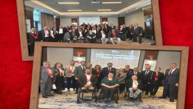 Kırmızı zemine yerleştirilmiş iki çerçeveli fotoğraf görünüyor. Üstteki karede geniş bir salonun içinde kalabalık bir grup birlikte poz veriyor; arka planda sunum perdesi açık. Alttaki karede yine aynı salonda, bu kez daha yakın planda bir grup fotoğrafı var. Ön sırada tekerlekli sandalyede oturan kişiler, arkalarında ise ayakta duran katılımcılar bulunuyor. Her iki fotoğraf da bir genel kurul toplantısının ardından çekilmiş hatıra kareleri izlenimi veriyor.