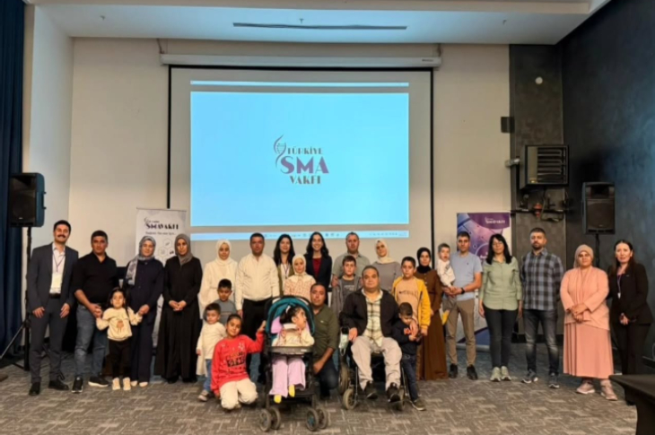 Bir konferans salonunda, sahne önünde bir grup yetişkin ve çocuk birlikte poz veriyor. Arkadaki ekranda “Türkiye SMA Vakfı” logosu yer alıyor.