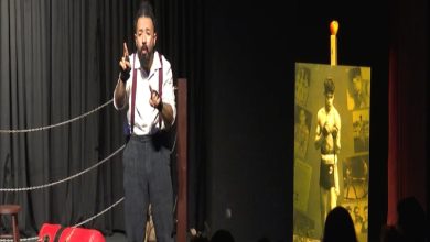 Sahnede tek başına duran bir oyuncu, işaret diliyle performans seriliyor gibi görünüyor. Arka planda boks ringini andıran halatlar, yerde kırmızı bir boks kum torbası ve yanında eski bir boksörün fotoğraflarının yer aldığı sarı tonlu bir pano bulunuyor. Seyirciler karanlıkta, ön planda siluet olarak seçiliyor.