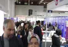 Görselde kalabalık bir fuar alanı görülüyor. Salon içinde birçok stant yer almakta ve insanlar bu stantlarda bilgi alıyor ya da sohbet ediyor. Bazı kişiler ellerinde broşürler taşıyor, bazıları ise oturup görüşme yapıyor. Üst kısımda “EXIT” tabelası dikkat çekiyor. Ortam hareketli ve sosyal bir etkinlik havasında; fuar, kariyer günü ya da tanıtım organizasyonu izlenimi veriyor.