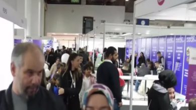 Görselde kalabalık bir fuar alanı görülüyor. Salon içinde birçok stant yer almakta ve insanlar bu stantlarda bilgi alıyor ya da sohbet ediyor. Bazı kişiler ellerinde broşürler taşıyor, bazıları ise oturup görüşme yapıyor. Üst kısımda “EXIT” tabelası dikkat çekiyor. Ortam hareketli ve sosyal bir etkinlik havasında; fuar, kariyer günü ya da tanıtım organizasyonu izlenimi veriyor.