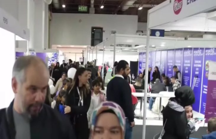 Görselde kalabalık bir fuar alanı görülüyor. Salon içinde birçok stant yer almakta ve insanlar bu stantlarda bilgi alıyor ya da sohbet ediyor. Bazı kişiler ellerinde broşürler taşıyor, bazıları ise oturup görüşme yapıyor. Üst kısımda “EXIT” tabelası dikkat çekiyor. Ortam hareketli ve sosyal bir etkinlik havasında; fuar, kariyer günü ya da tanıtım organizasyonu izlenimi veriyor.