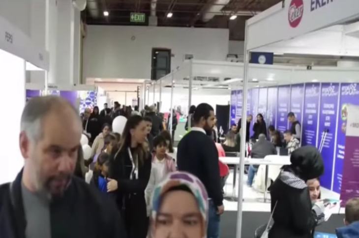 Görselde kalabalık bir fuar alanı görülüyor. Salon içinde birçok stant yer almakta ve insanlar bu stantlarda bilgi alıyor ya da sohbet ediyor. Bazı kişiler ellerinde broşürler taşıyor, bazıları ise oturup görüşme yapıyor. Üst kısımda “EXIT” tabelası dikkat çekiyor. Ortam hareketli ve sosyal bir etkinlik havasında; fuar, kariyer günü ya da tanıtım organizasyonu izlenimi veriyor.