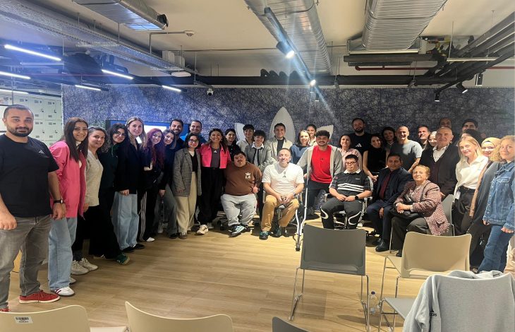 Görselde, geniş bir grup bir araya gelmiş şekilde poz veriyor. Farklı yaş ve özelliklerde birçok kişi ayakta veya sandalyelerde oturarak gülümsüyor. Ortam modern bir iç mekân; tavan aydınlatmaları, havalandırma boruları ve ahşap zemin dikkat çekiyor. Genel atmosfer sıcak, kapsayıcı ve topluluk ruhunu yansıtan bir buluşma izlenimi veriyor.