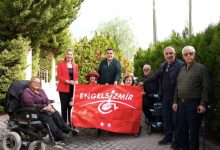 Yeşillikler içerisinde bir alanda yerel yöneticiler, belediye personeli ve engelli bireyler, “Engelsiz İzmir” yazılı kırmızı bayrağı birlikte tutarak gülümseyen yüzlerle poz veriyor.