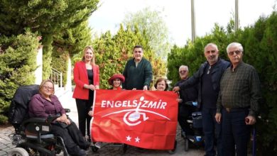 Yeşillikler içerisinde bir alanda yerel yöneticiler, belediye personeli ve engelli bireyler, “Engelsiz İzmir” yazılı kırmızı bayrağı birlikte tutarak gülümseyen yüzlerle poz veriyor.
