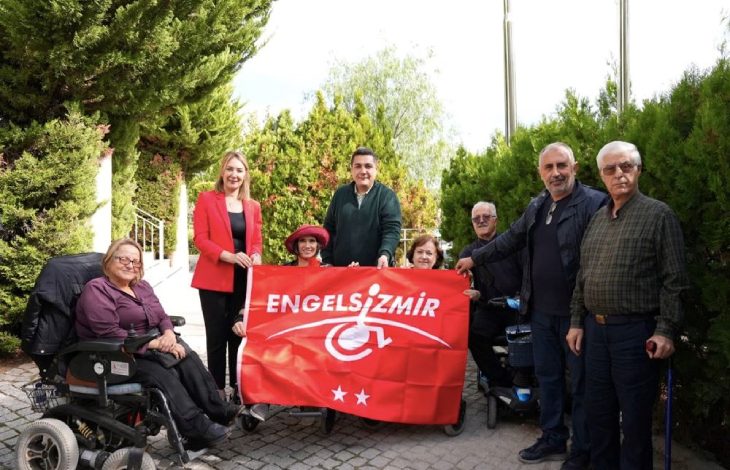 Yeşillikler içerisinde bir alanda yerel yöneticiler, belediye personeli ve engelli bireyler, “Engelsiz İzmir” yazılı kırmızı bayrağı birlikte tutarak gülümseyen yüzlerle poz veriyor.