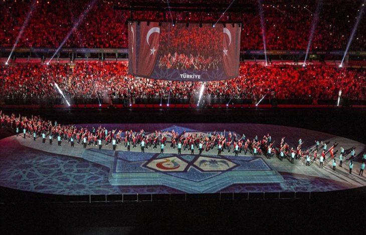Büyük bir stadyumda gerçekleştirilen görkemli bir tören görülüyor; sahnenin ortasında ışık gösterileri ve semboller yer alırken, sahada Türk bayrakları taşıyan sporcular yürüyüş yapıyor ve tribünlerde kırmızı ışıklarla aydınlatılmış kalabalık bir izleyici topluluğu bulunuyor.
