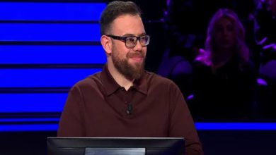 Bir televizyon stüdyosunda yarışma kürsüsünün arkasında duran bir erkek görülüyor. Gözlük takıyor, sakallı ve sakin bir ifadeyle sahneye odaklanmış. Mavi tonlu ışıklar ve arka plandaki seyirci siluetleri, yarışma atmosferini belirginleştiriyor.