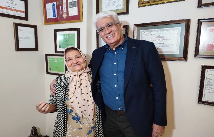 Duvarı sertifika ve plaketlerle dolu bir odada, yaşlı bir kadın ile yanında duran bir adam yan yana gülümseyerek poz veriyor. Kadın başörtülü, çiçek desenli elbise ve hırka giyiyor; adam ise koyu renk bir ceket ve gömlekle şık bir görünüme sahip.