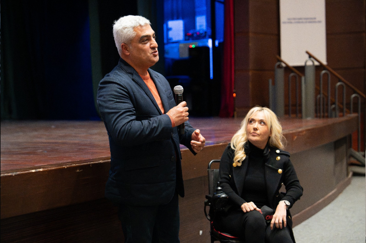 Bir salonun ön kısmında, sahnenin kenarında iki kişi yer alıyor. Ayakta duran erkek mikrofonla konuşurken, yanında tekerlekli sandalyede oturan kadın onu dikkatle dinliyor. Ortam ciddi ama sıcak; ışıklar konuşmanın önemini vurguluyor. Arka planda sahne, basamaklar ve salonun iç düzeni seçiliyor.