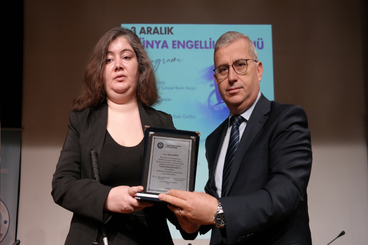 Bir sahnede iki kişi yan yana durarak bir plaket tutuyor. Arkadaki ekranda “3 Aralık Dünya Engelliler Günü” başlıklı program görünüyor. Her iki kişi de resmi kıyafetler içinde, törensel ve ciddi bir atmosferde kameraya poz veriyor.