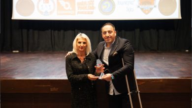 Sahnenin önünde iki kişi yan yana duruyor; ikisi de kameraya dönük ve ellerinde birlikte tuttukları bir ödül bulunuyor. Arkada çeşitli kurum logolarının yer aldığı büyük bir projeksiyon perdesi var. Ortam resmî, tören havasında; sahne ışıkları ödül anının önemini vurguluyor.